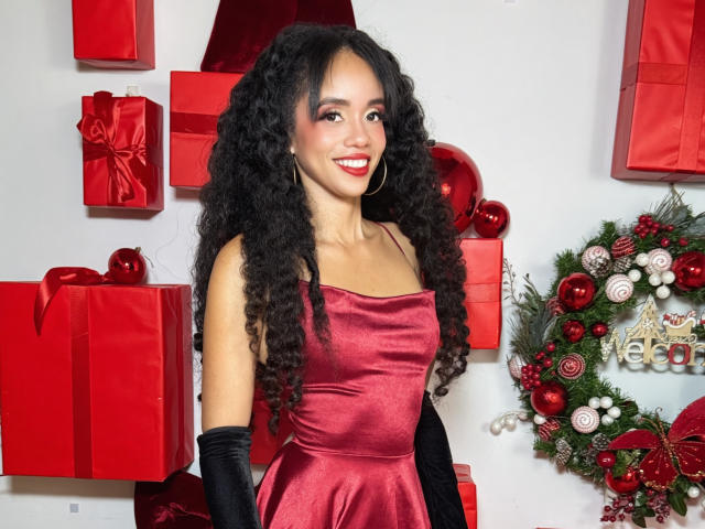 TerryGreen with curly black hair, wearing a red satin dress, black gloves, and red lipstick, smiling, standing in front of red gifts and a Christmas wreath.
