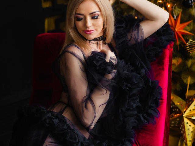 MiladyOne with blonde hair, black sheer dress, and black lace choker, sitting on red velvet chair, black ruffled fabric covering her tits, arm raised, sexy, sultry look.