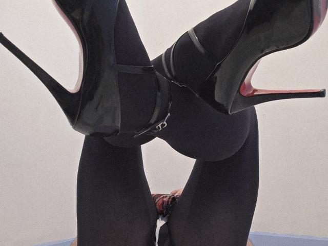SissyBetty's black glossy heels and tight black pants, with her ass up and legs spread, showing a thong and a small butt plug in her butt.