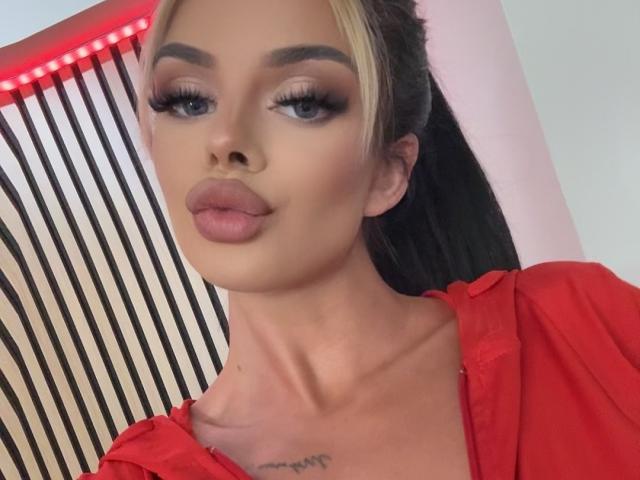 IvyRaven has blonde and black hair, heavy eyeliner, long lashes, pink lips, a red shirt, and a tattoo on her chest. She's looking at the camera with a pout.