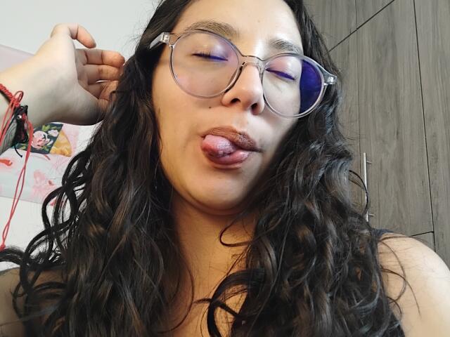 Mllevv has long, wavy black hair, purple-tinted glasses, and is sticking out her tongue with her lips slightly parted. She's holding her hair with one hand.