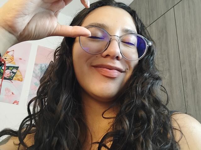 Mllevv with long wavy black hair, glasses, and pink tongue out, touching nose with finger, smiling. Light brown skin, wearing black top, wooden wall in background.