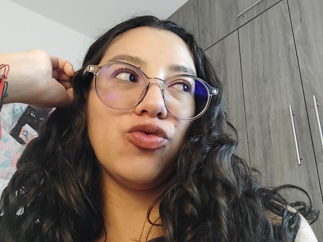 Mllevv has long black wavy hair, clear glasses, and is pouting with slightly parted pinkish-brown lips, looking to the side with a red bracelet on her wrist.