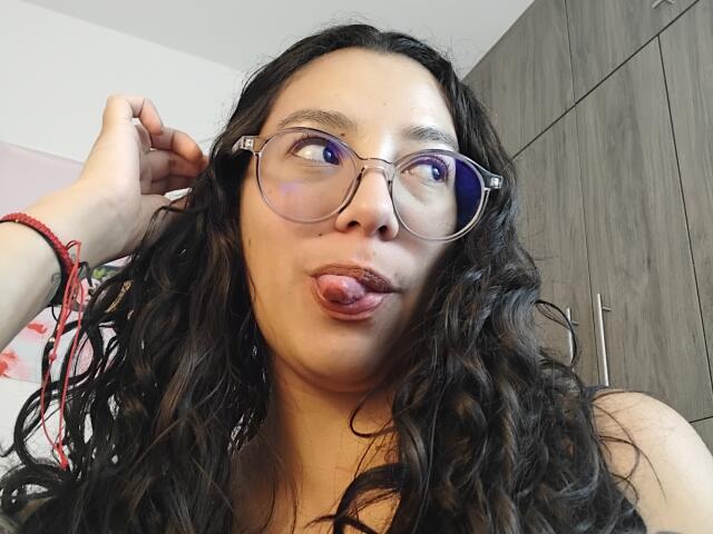 Mllevv has long, wavy black hair, glasses, and is sticking out her tongue. She's wearing a red bracelet and has her hand in her hair.