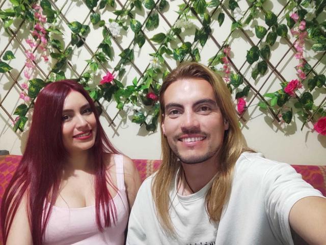 JackyEmma with long red hair and fair skin is on the left, wearing a pink tank top. JackyEmma has a slim build and a slight smile. JackyEmma on the right has long blonde hair, a fair complexion, and a mustache. He is wearing a white t-shirt and is smiling directly at the camera. Both JackyEmmas are sitting on a pink patterned couch. Green vines with pink flowers are arranged in a diamond pattern on the wall behind them. The image is a selfie with both JackyEmmas in the frame.