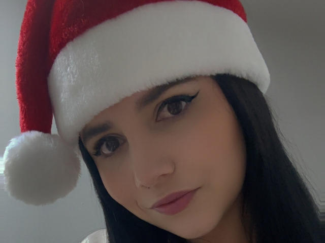 Kkatalina with dark hair, wearing a red Santa hat with white trim, has brown eyes, black eyeliner, and pink lips, in a soft light. Her nose is pierced.
