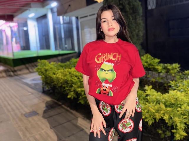 IrisBlair with long dark hair, wearing a red "Grinch" t-shirt and black pants with pizza prints, standing outside at night, hands near her ass.