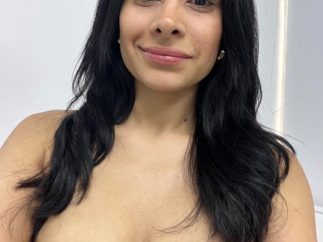 LyraRaven with black wavy hair, medium brown skin, and pink lips, smiling slightly, showing small nipples and bare shoulders, wearing small gold earrings, in a selfie.