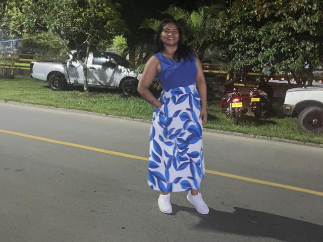 SusanaDiaz stands on a street, smiling, wearing a blue sleeveless top and white-blue floral skirt with white sneakers, hands in pockets, dark hair, medium brown skin.