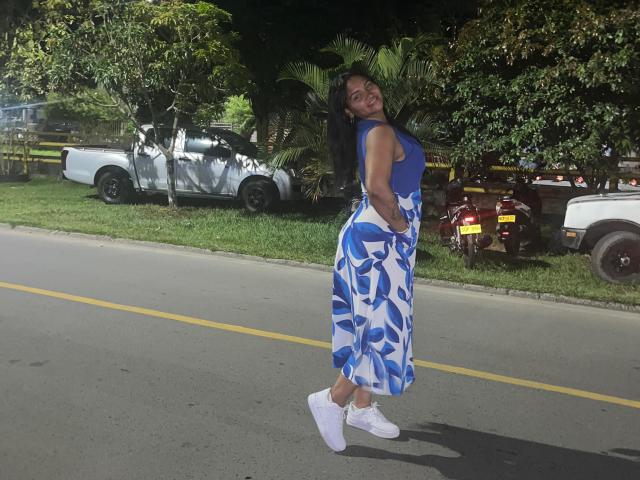 SusanaDiaz with blue sleeveless top, white and blue floral skirt, white socks, and white sneakers, standing on road, looking back, hands in skirt, dark hair, daytime.
