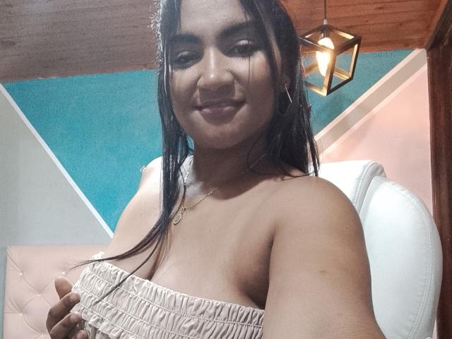 SusanaDiaz with wet black hair, smiling, wearing a strapless beige top, showing her small tits, holding the top, in a room with a wooden ceiling and hanging light.