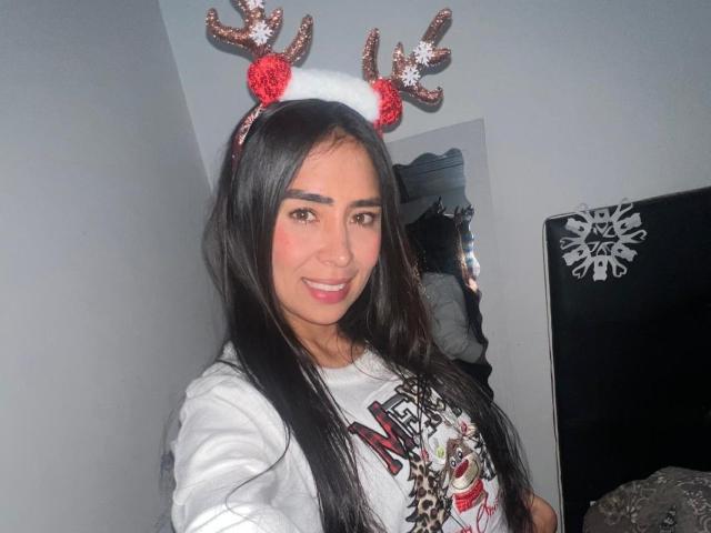 AngelinaLina with long black hair, wearing sparkly red and white reindeer antlers, smiling at the camera, in a white graphic tee, with her tits subtly visible.