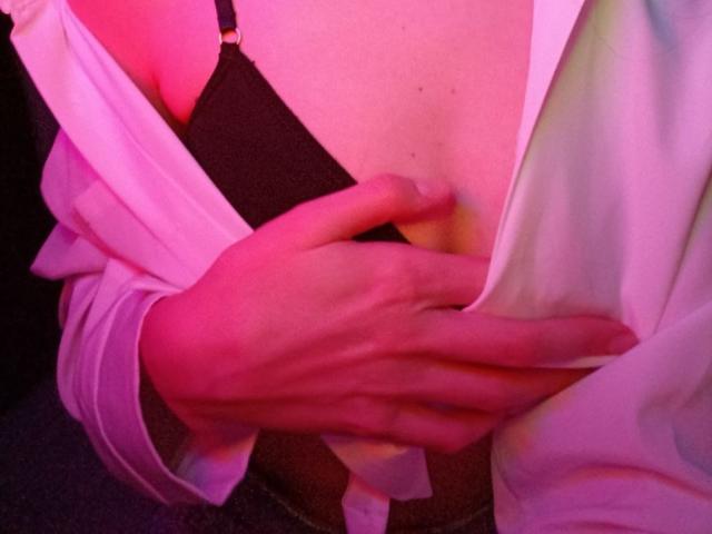 WollyMolly's hand is gently holding their bare chest, exposing a black bra with a gold clasp, under pink and white fabric. Visible are WollyMolly's small, bare tits.