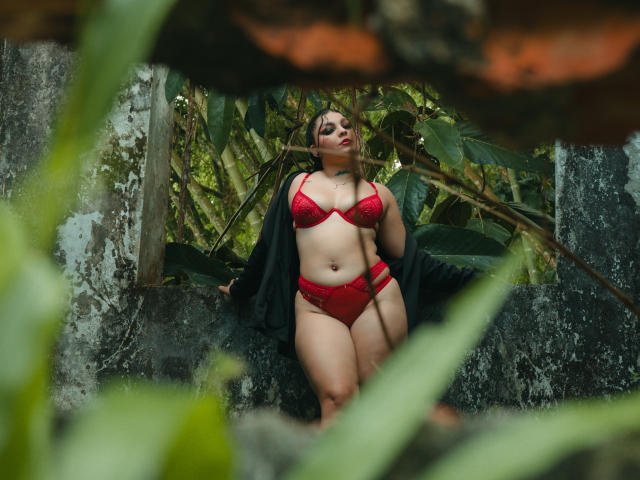 MelodyBlossom stands in lush greenery, wearing red lace lingerie with matching panties, black jacket open, showing her curvy, pale-skinned body, small tits, and full ass.