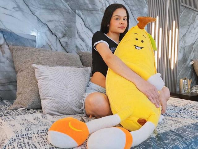 MiaGalvez with small tits, wearing a black shirt and denim shorts, sits on a bed hugging a yellow banana plushie with orange ears and white socks.