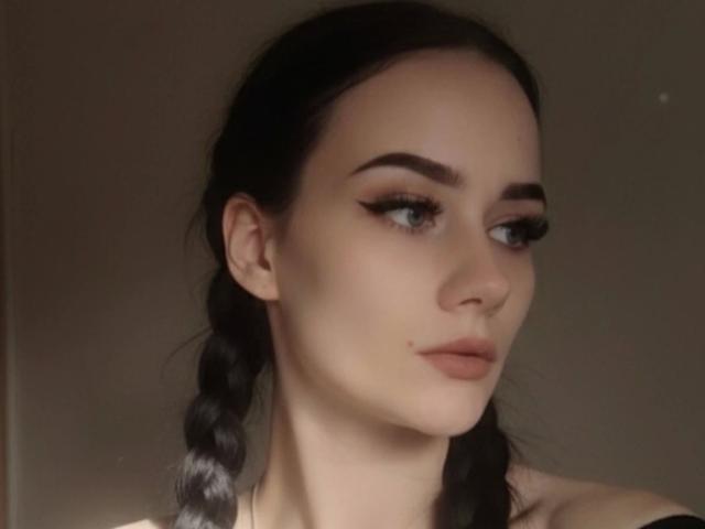 NinaLecker-hot has pale skin, dark eyebrows, long black eyelashes, and pouty pink lips. She has black braids and a small beauty mark on her right cheek.