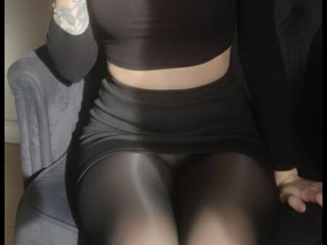 NinaLecker-hot's black crop top and tight skirt with shiny black pantyhose, sitting on a blue chair, tattoo on her right arm, black jacket partially visible.