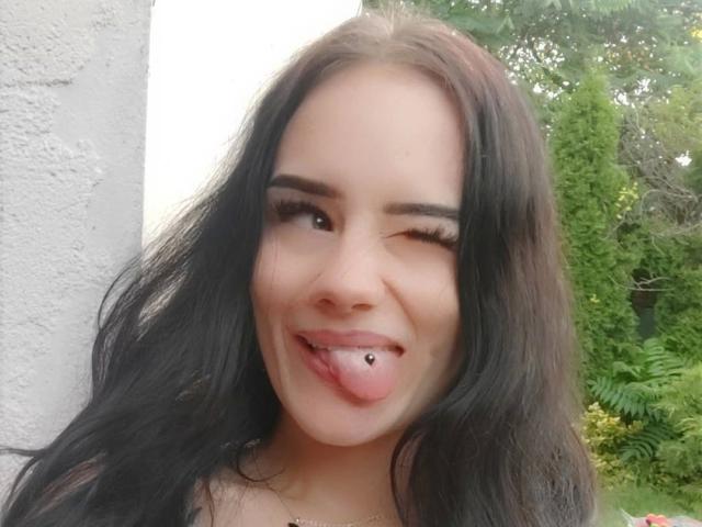 NinaLecker-hot with long black hair, winking, sticking out her tongue with a pierced lower lip, wearing eyeliner and a necklace, against a white wall.