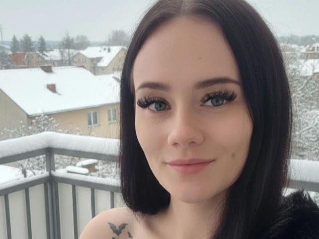 NinaLecker-hot has black shoulder-length hair, blue eyes, and thick black eyelashes. She's smiling with a small mole on her left cheek, wearing a black top with a bird tattoo.