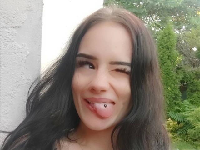 NinaLecker-hot with long black hair, winking, sticking out her tongue with a small lip piercing, standing near a white wall.