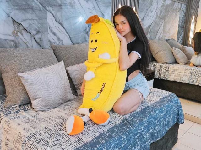 MiaGalvez with long black hair, cute face, and small tits, sitting on a bed, hugging a yellow banana stuffed animal with a smiling face, wearing a black top and denim shorts.