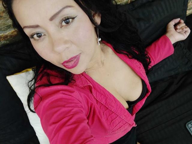 ThaliaSaenz with black hair, pink jacket, and pink lipstick, showing her cleavage in a black bra, lying on a black pillow, smiling at the camera.