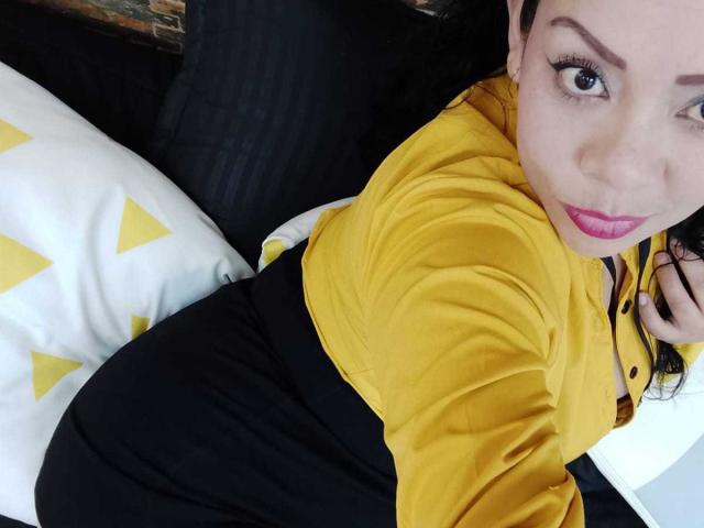 ThaliaSaenz with yellow shirt, black skirt, pink lipstick, and black hair, lying on black bed, white pillow with yellow triangles, looking up at camera with blue eyes.