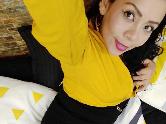 ThaliaSaenz with yellow top, black skirt, and pink lipstick, holding her right boob, smiling, black wavy hair, stone wall in background, white and yellow pillow.