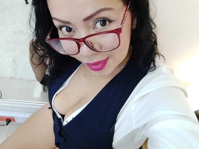 ThaliaSaenz with red glasses, wavy black hair, pink lipstick, deep cleavage in a navy blue vest, white shirt, and white bra, smiling at the camera.