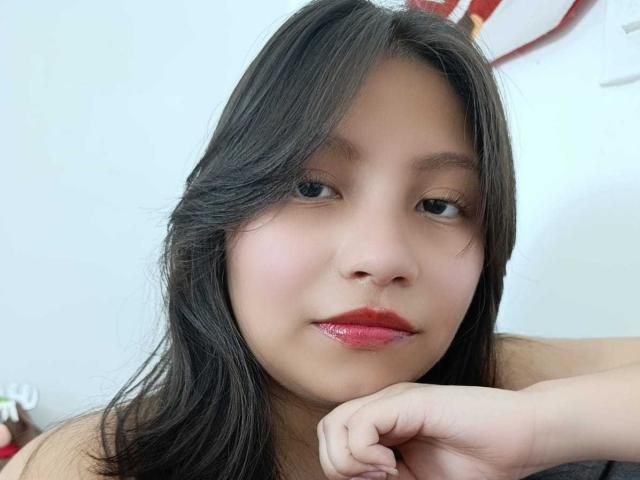 NovaSweet has shiny pink lips, black straight hair, and a soft smile, resting her chin on her hand. Her skin is light, and she's looking to the side.