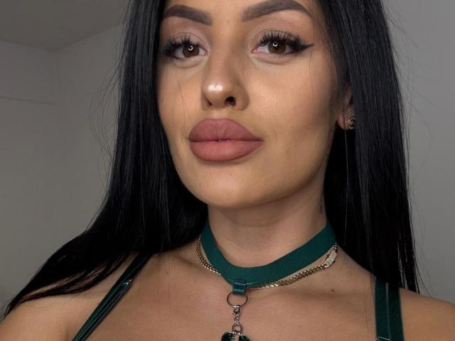 BoobsiEmilaa has long black hair, brown eyes, and pouty pink lips. She's wearing a green collar with a chain and has a green strap over her tits.