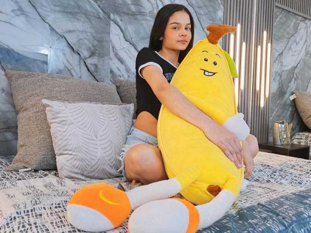 MiaGalvez, with long dark hair, sits on a bed hugging a yellow, cartoonish, smiling stuffed toy with orange and white shoes. She wears a black top and denim shorts.