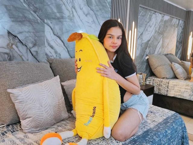 MiaGalvez with long brown hair, black shirt, and denim shorts, hugs a yellow plush banana with a cute face on a gray-patterned bed, her small ass slightly visible.