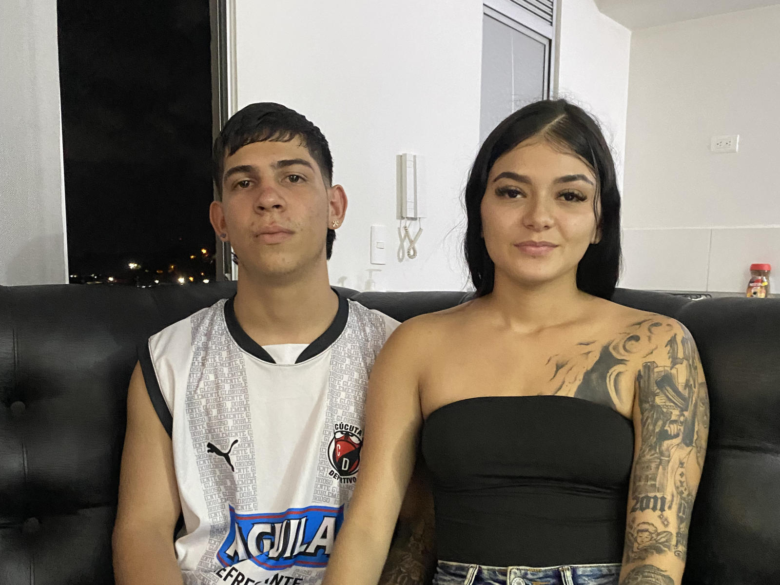 FlacaHermosax with short brown hair, wearing a white Puma sports shirt, sits next to FlacaHermosax with black strap top, visible tattoo on right arm, in a black leather sofa.
