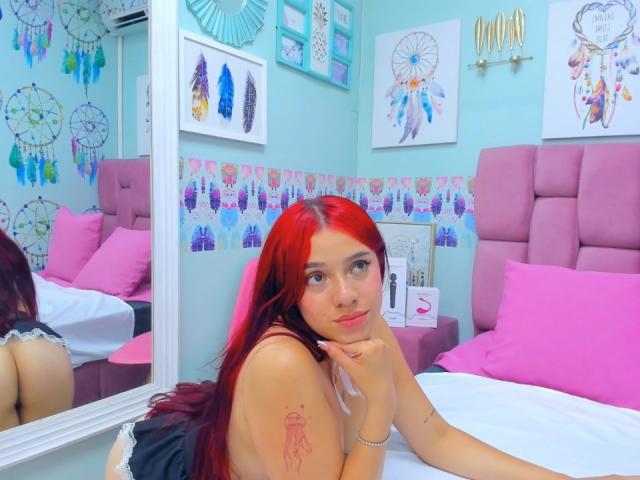 MiaLeroy, con i capelli rossi, piccoli seni, un tatuaggio sul braccio, sdraiata sul letto, con cuscini rosa, riflesso nello specchio, che guarda la fotocamera, con la mano sul mento, carina, pelle chiara, occhi azzurri, camera da letto..