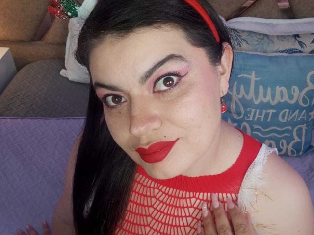 Gaiax69 with dark hair, red lipstick, and red fishnet top, wearing red headband, looking at camera, showing small nose, freckles, and red earrings.