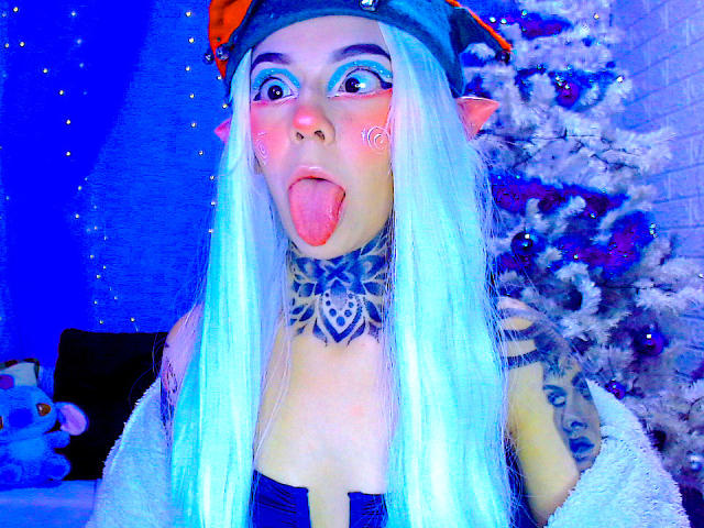 LannieGonzales has long white hair, elf ears, blue eye makeup, and a blue choker with a mandala pattern. She's sticking her tongue out, showing off tattoos and a nose ring.