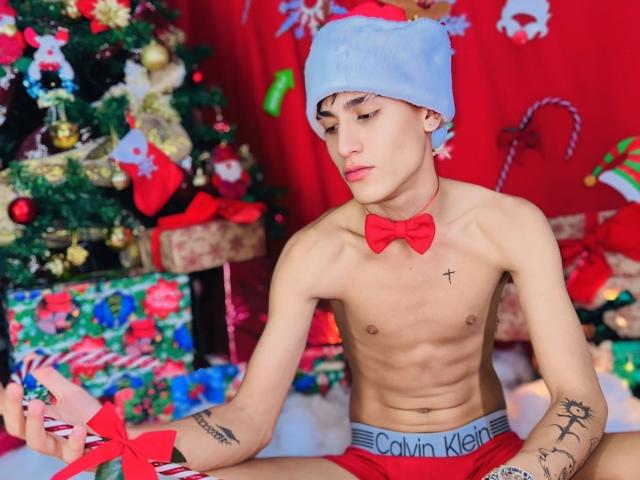JustXJake, shirtless with a red bowtie and blue Santa hat, holding a red ribbon, has tattoos, is wearing Calvin Klein underwear, and is sitting in front of a Christmas tree.
