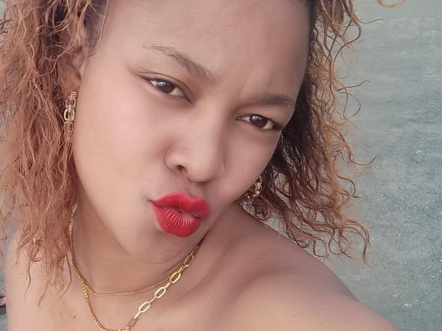 Faranys with red lipstick, curly brown hair, gold earrings, and gold chain, puckering her lips in a selfie, showing her bare shoulders and a slight frown.