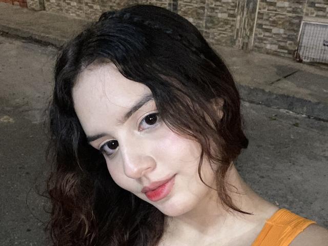 KylieLumi has pale skin, dark wavy hair, and small tits, with light makeup and red lips, wearing an orange top, smiling softly, and looking at the camera.