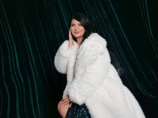 MoonVelour with a fluffy white coat, dark hair, and a subtle smile, touching her cheek. She's wearing sequined blue pants and has a cute, confident look.
