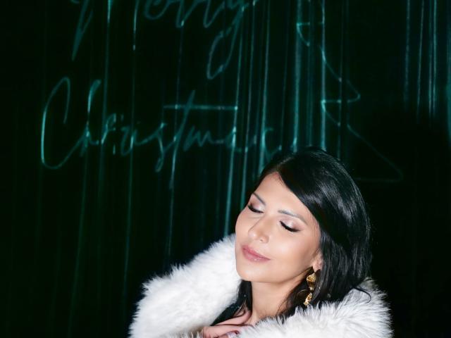 MoonVelour with dark hair, closed eyes, and pink lipstick, wearing a white fur collar, gold earrings, and red nails, smiling softly.