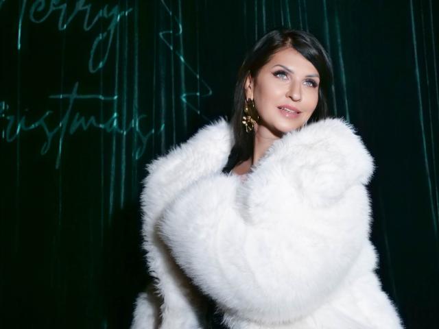 MoonVelour with pale skin, blue eyes, and dark brown hair, wearing a fluffy white fur coat, holding it close to her chest, smiling slightly, wearing gold star-shaped earrings.