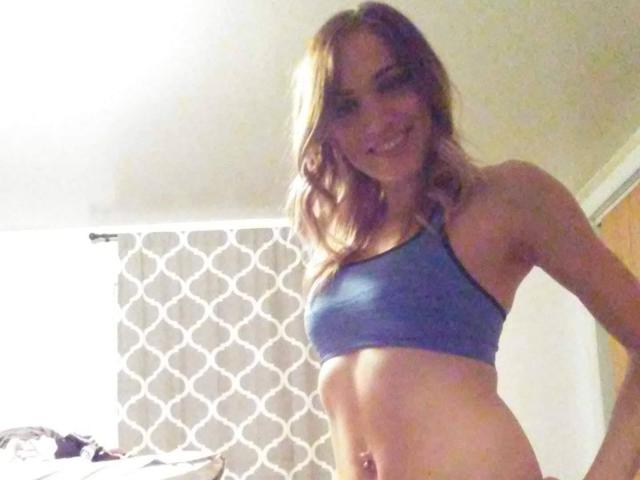 PhoenixPaulline with blonde hair, blue sports bra, and toned abs, smiling in a bedroom. Her tits are small, and her belly button is pierced.