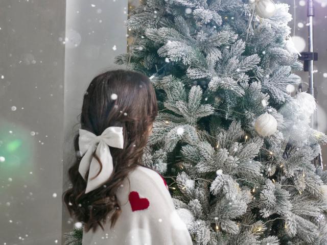 OwnPath with brown hair in a white bow, facing a frosted Christmas tree with white and red ornaments, wearing a white sweater with red hearts. Snowflakes are visible.