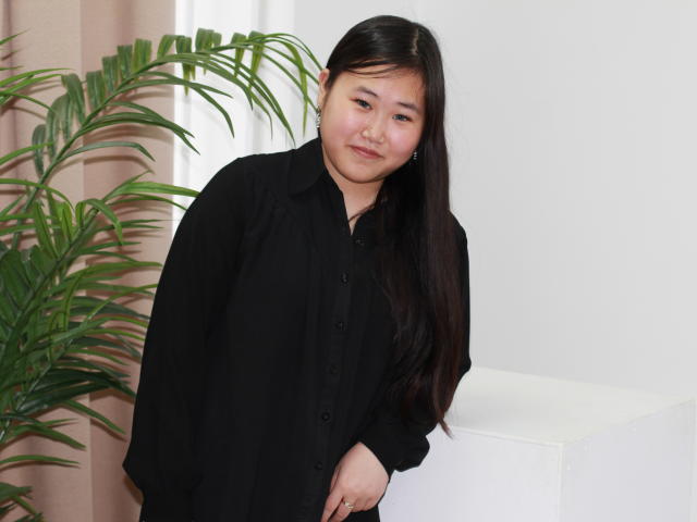 AnoraOliver, Asian woman with long black hair, wearing a black button-up shirt, smiling slightly, standing next to a green plant, white wall, and a white cube.
