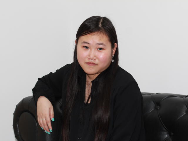 AnoraOliver with long black hair, wearing a black shirt, sitting on a black leather couch, arm resting on the back, turquoise nail polish, white background.