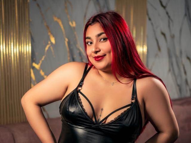 ZoeEvansl with red hair, black leather lingerie, and red lipstick, smiling at the camera, hands on hips, showing her tits and a small cross necklace.