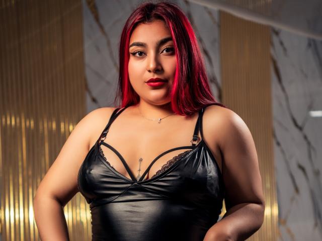 ZoeEvansl with red hair, wearing a black leather lingerie top, showing her medium-sized tits with lace straps, standing with hands on hips, looking at camera.