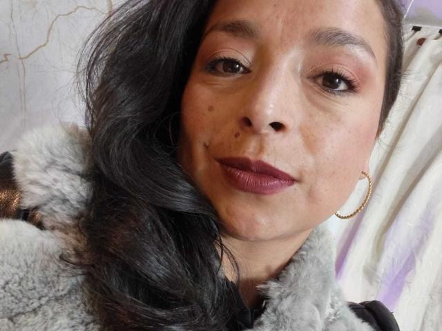 StangelarRivera has dark hair, brown eyes, pinkish eyeshadow, dark lipstick, a small mole on the left side of her nose, and a gold hoop earring, wearing a furry grey coat.